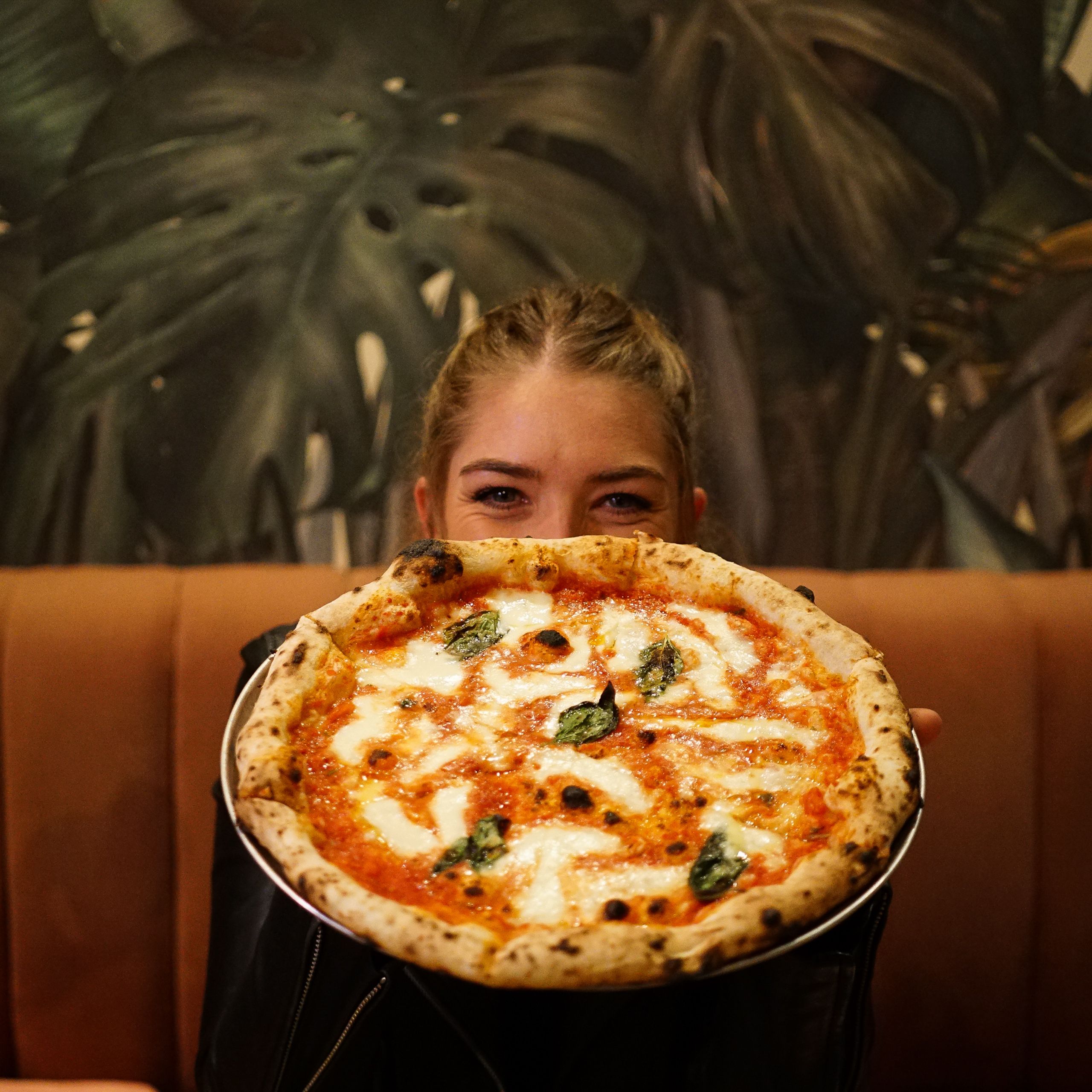 Mulher segurando uma pizza diante do rosto, com parede de folhagens pintadas ao fundo.