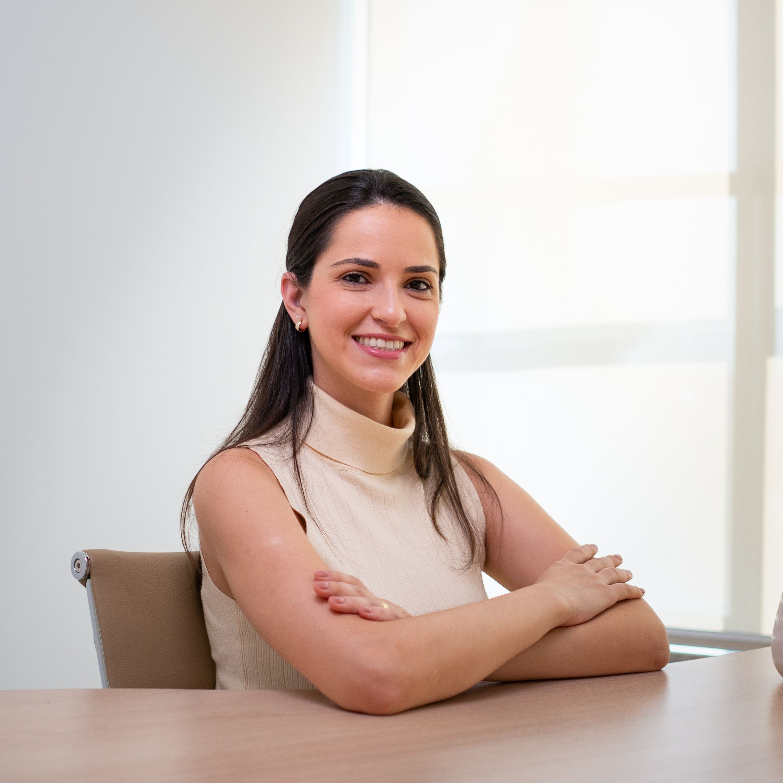 Retrato profissional de fonoaudióloga sentada à mesa, de braços cruzados, em consultório de parede clara.